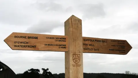 A wooden fingerpost with Bourne Bridge, Ipswich Waterfront, Suffolk Food Hall, Pin Mill and Shotley Gate written on it. In the middle there is also the St Joseph's College emblem.