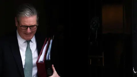 Reuters Sir Keir Starmer in a dark suit with a white shirt and green tie and carrying two folders walks out of 10 Downing Street. 