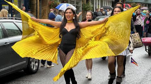 A drag queen walks down the parade in a black corset spreading golden wings