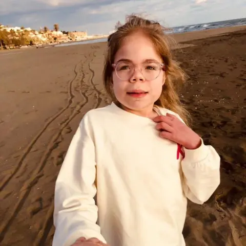 Darren Scott Sophia Scott is standing on a beach. She is a young girl with long, blonde hair and is wearing large, pink-rimmed glasses. She is wearing a cream-coloured sweatshirt and half smiling at the camera.