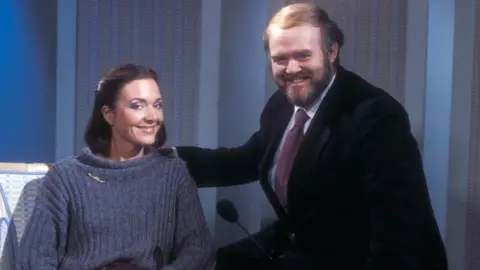 Anna Ford, with brown hair and a grey jumper, sits next to Donnie B MacLeod, a large bearded man in a dark jacket with burgundy tie and blue and red checked shirt. They are both smiling broadly.