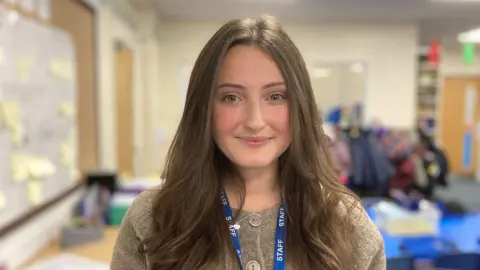 Year 1 teacher Emma Westmoreland is smiling for the picture. She has long brown hair and is wearing her staff ID lanyard. She is standing in her classroom. 