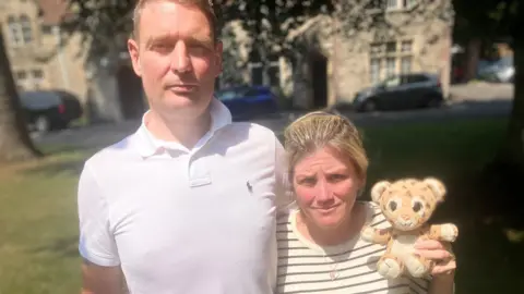 Barry and Michelle Curtis. Barry has short hair and is wearing a white polo shirt. He is stood next to Michelle and has his arm around her. Michelle has blonde hair that's tied up and is wearing a striped t-shirt. She is holding a small leopard teddy bear.