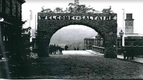 An old black and white photo shows a huge decorative arch made of flora and foliage, with the word 'Saltaire' across the top in large letters. It sits in a cobbled street, and mill style buildings and a tower in the background in indicate this is an industrial area. Figures in the background wear bowler hats and clothing which looks late 19th Century in style.