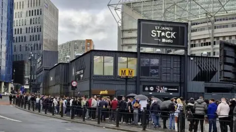 A queue of people standing outside St James' Stack which is made up of black shipping containers.