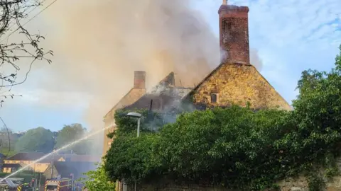 Stone building with fire engines parked in front and water from a firefighter's hose spraying onto the building.