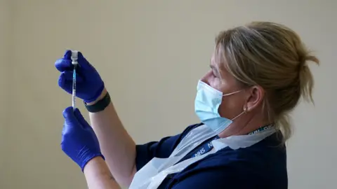 A blonde nurse with protective coverings on her face, hands and chest holds up a needle and vaccine vial.