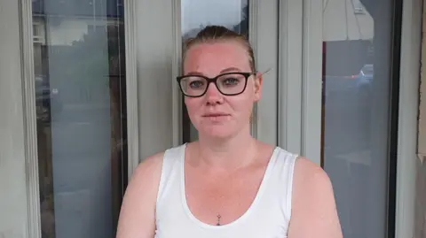 Roberta, a woman with light brown hair tied back from her face, is looking at the camera. She is wearing a white vest top and black framed glasses.