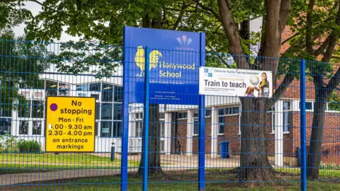 A blue sign that says "Honywood School" in a yellow font, with a yellow cockerel also on it. The school building is in the background and is a mixture of orange brick and large white walls with windows.