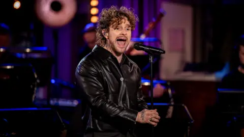 Tom Grennan pictured singing in a dimly lit studio. He is wearing a black leather jacket and has curly dark blonde hair. He is holding a microphone stand and has tattoos on his hand.