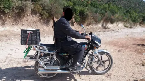 Met Police Mohammad's nephew on a motorbike. His face has been obscured but he is wearing a black outfit with black shoes, and he has black hair. The motorbike is also black with silver features and has a plastic box on the back. The picture has been taken on a dirt track in front of lots of bushes and trees.