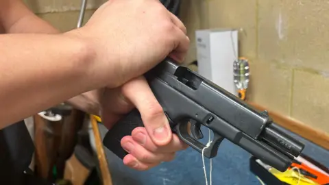 Two hands are seen cocking a black semi-automatic pistol-style gun