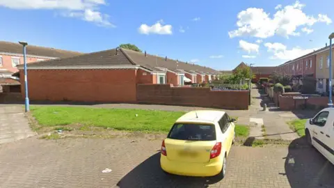 A row of red brick bungalows with a yellow car parked in a small car park next to them. They are pictured from a gable end, stretching out into the background. The homes are built between and parallel to two rows of two-storey terraced houses. A footpath runs between the different rows.