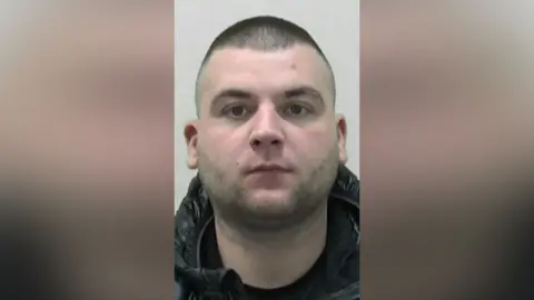 A mugshot of Connor Wade. He has a round head with faint dark stubble and cropped black hair. He is wearing what appears to be a black outdoor jacket.