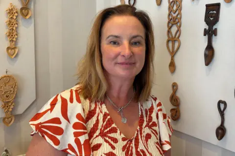 Jane, who has light brown hair and wears a floral white and red top, with love spoons attached to a wall in the background. 