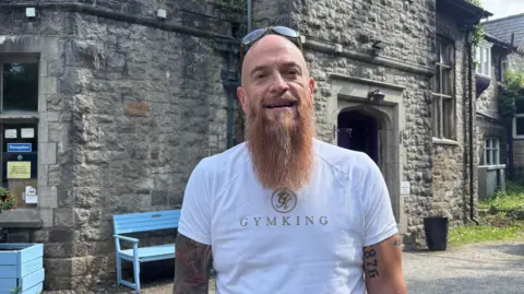 James Deakin stands outside historic large grey brick building with a light blue bench in front of it. He is bald with a long beard with sunglasses on his head and has a sleeve of tattoos.