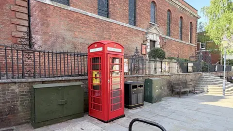 A red telephone box labelled “defibrillator” stands on a pavement outside a brick church building in Guildford, with benches, bins and street fixtures nearby.