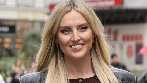 Getty Images Perrie Edwards pictured out walking in London. Perrie has long blonde hair worn loose and blue eyes. She wears a black leather jacket over a brown top. 