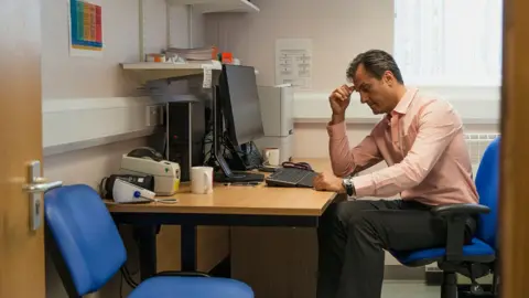 Getty Images A GP sits at his desk next to an empty patients chair in his surgery, seen through an open door. He is wearing a shirt and dark trousers and is looking thoughtful.