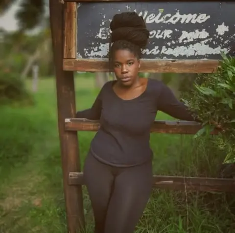 Mamkhabela Mthembu Mamkhabela is a young black woman. She wears a black, long-sleeved top and black leggings. Her braids are piled on top of her head in a large bun, and she stares directly into the camera.