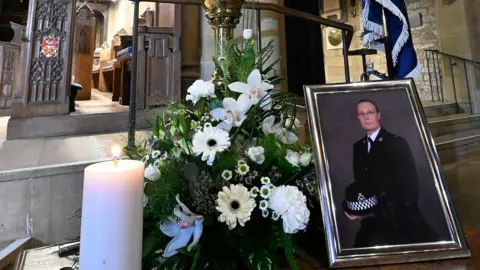 West Yorkshire Police A framed photo of a police officer in uniform, next to flowers and a lit candle. The items are in the front of a cathedral.