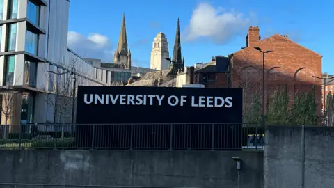 A black sign with University of Leeds. The sign is behind a railing. It is next to a modern building and in the background there is the University tower. It is a sunny day.