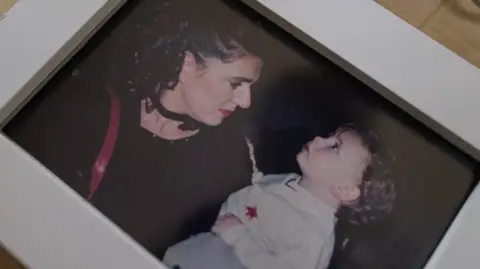 Geraldine looks into the eyes of her child while wearing a black top and black necklace. She has black curly hair which has been tied up and is wearing pink lipstick. Her child has brown curly hair and is wearing a white jumper. 
