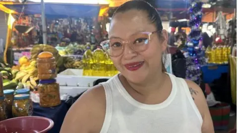 Nciole Castillo speaks to the BBC from her market stall in Tegucigalpa. She has her hair tied back and is wearing glasses and a white tank top. She is smiling into the camera. 
