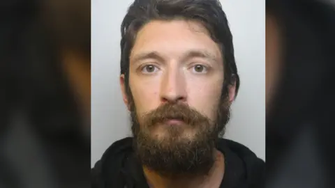 Avon and Somerset Police A police custody photo of David Parish. He has short dark brown hair and a thick dark brown beard. He is wearing a black hooded jumper.