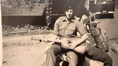 Handout Mr Peace holds a guitar and looks at the camera. He is wearing a shirt and trousers and a black beret-style cap in the black and white photo.