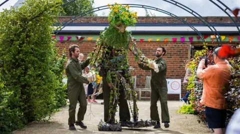 Two men in green overalls stand either side of, and manipulate a larger than life-size puppet with a frame covered with plants and flowers. They are standing in front of a brick building, with a bank of shrubs on either side. To the left a man in an orange shirt is taking a photograph.