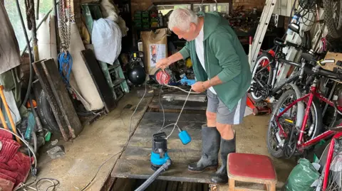 Kate Bradbrook/BBC Imre Berg, en su garaje, con una bomba de agua, botas Wellington, pantalones cortos, una camiseta verde y una camiseta blanca. En su garaje hay muchos objetos, incluyendo herramientas y bicicletas.