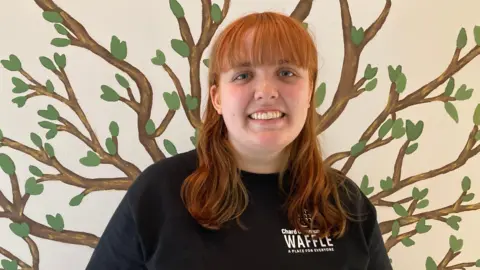 Manager Bleu Gover is pictured smiling into the camera. She is wearing a black jumper with Chard CommuniTEA cafe written on it. She is standing in front of a tree painting on a wall.