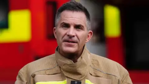 PA Media A man with dark brown and grey hair. he is wearing a light brown fire suit wich has yellow strips on the breast