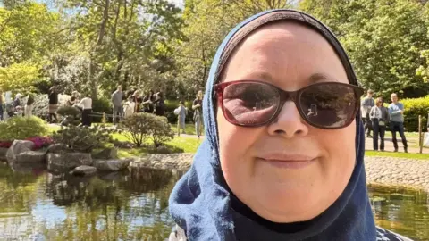 Kimberly Lavery Kimberly, a woman wearing a blue headscarf and sunglasses, is smiling for the camera. Behind her is a pond and people wandering past in a park.