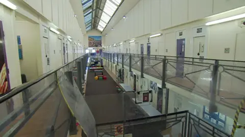 The image shows the interior of Wetherby Young Offender Institution viewed from an upper-level walkway. Two tiers of doors line both sides of a long, corridor, each door fitted with observation panels and secure locking mechanisms. The ceiling is high and fitted with large skylights. On the ground floor there is recreational or gym equipment, along with posters and noticeboards on the walls. 