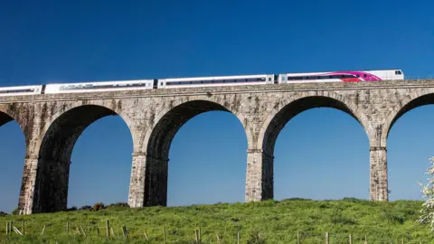 A bridge, on top of a field of grass, with a train crossing it. There is a blue sky in the background. 