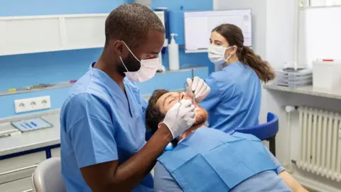 Getty Images Male dentist examines the teeth of a male patient who is lying back the chair with his mouth open. A female assistant sits in the background.