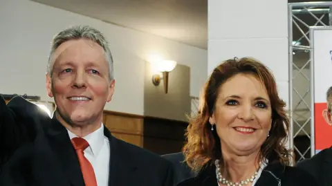 Pacemaker Peter and Iris Robinson at a DUP annual conference. He is wearing a dark suit with a white shirt and red tie. Iris is wearing a black suit with a pearl necklace.