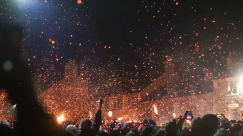 Jonathan Atkinson Photography Bright orange embers dot the night sky as a crowd of people, in silhouette, look up. Some are taking pictures on their smartphones. 