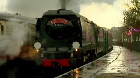A steam train arrives at a platform, decorated with coloured lights, as steam comes out of the funnel.