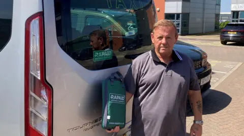 Billy Hughes, standing by a large grey taxi, holding a green kitbag with a zip branded with the word Rapaid