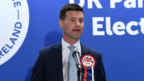 PA Media Jonathan Buckley has short black hair and is wearing a dark suit, white shirt and light blue tie and a DUP election rosette. He is wearing a microphone