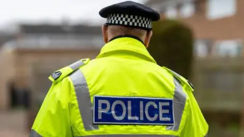 A policeman is seen from behind. He is wearing a black hat with a black and white checkered band, and a hi-vis yellow jacket bearing the word 'police' on his back. 