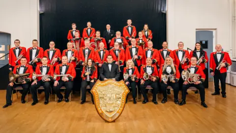 Members of a brass band wearing red jackets, bow ties and holding instruments, sit behind a big golden shield in a town hall.