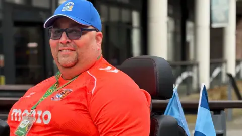 A smiling Craig Wyatt is wearing a bright orange Coventry City shirt and blue cap. He is sitting on a mobility scooter with Coventry City flags on the rear