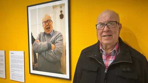A man in his 80's smiles in front of a photo of himself at an exhibition