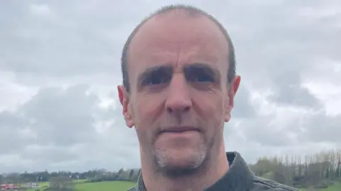 Mark H Durkan, in green shirt, with grass and trees in the background.