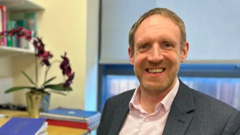 Prof John-Paul Taylor is seated in his office at Newcastle University's Centre for Ageing and Vitality. He is wearing a grey suit and pink shirt. He has brown hair and stubble. Behind him are different coloured exercise books on his desk and books on a shelf.