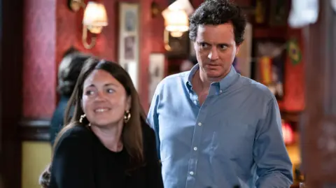 Will Ellis as Theo Hawthorne in EastEnders, standing next to character Stacey Slater, in the Queen Vic